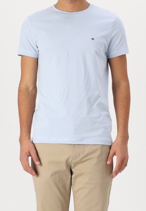 Man draagt lichtblauw T-shirt met korte mouwen en klein logo op de borst, gecombineerd met beige broek, staand tegen een effen achtergrond.