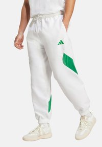 Bijele trenirke od sjajnog materijala s zelenim detaljima, s elastičnom gumicom u pojasu, bočnim džepovima i suženim manžetama. Vidljiv Adidas logo.