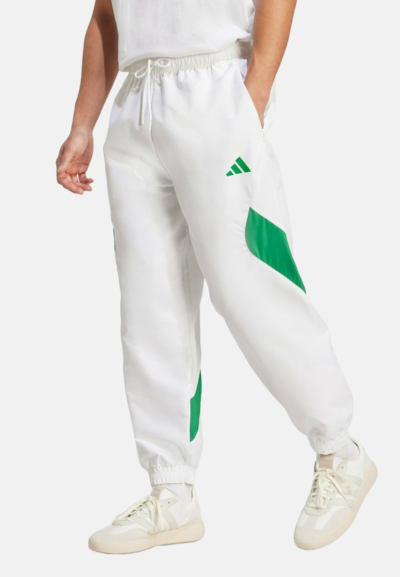 Bijele trenirke od sjajnog materijala s zelenim detaljima, s elastičnom gumicom u pojasu, bočnim džepovima i suženim manžetama. Vidljiv Adidas logo.