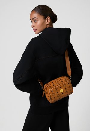 Donna che indossa un felpa nera e pantaloni, con una borsa a tracolla marrone a motivo con hardware dorato sulla spalla, che guarda di lato.
