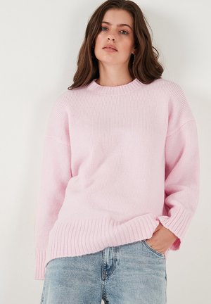 Light pink knitted sweater with a ribbed round neckline, dropped shoulders, and ribbed hem. Worn over blue denim jeans.