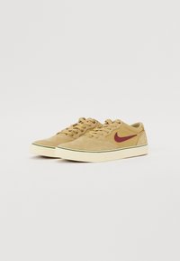 Beige Wildlederschuhe mit einem maroonfarbenen Nike-Swoosh, runden Schnürsenkeln und einer weißen Sohle, die eine grüne Akzentlinie um die Sohle hat.