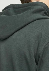 Kaptur w ciemnozielonym kolorze, z teksturowanej tkaniny, zbliżenie na kaptur i tył, prezentujące prosty, minimalistyczny design.