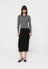 Femeie îmbrăcată cu un top gri cu mâneci lungi, o fustă midi neagră și sandale cu toc slingback negre, în fața unui fundal alb simplu.