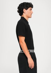 Cămașă polo neagră cu mâneci scurte, guler clasic. Pură cu pantaloni negri care au un brâu cu logo alb. Textură netedă, design ajustat.