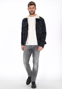 Chaqueta de cuadros negros y azules con un cuello de piel sintética beige, combinada con una camiseta blanca y vaqueros grises. Botas negras con cordones completan el conjunto.