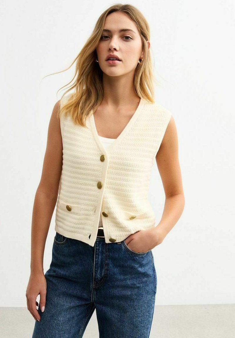 Strickweste in Creme mit V-Ausschnitt, fünf Vorderknöpfen und zwei Taschen, kombiniert mit hoch geschnittenen blauen Jeans. Glatte Textur, lässige Passform.