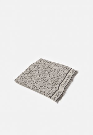 Beige en zwarte Calvin Klein-sjaal met herhalend "CK" logo patroon en franjes aan de randen, opgevouwen op een witte achtergrond.