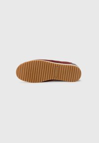 Suola di sneakers con scanalature orizzontali profonde e una etichetta logo rettangolare al centro su uno sfondo bianco semplice.