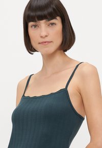 Camisole à côtes vert foncé avec de fines bretelles et une bordure en dentelle festonnée le long de l'encolure. Texture lisse avec une coupe ajustée.