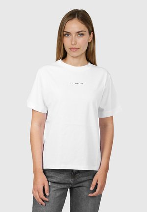 Junge Frau mit glatten hellbraunen Haaren, die ein weißes T-Shirt mit kleinem zentriertem Text und dunkelgraue Jeans trägt, vor einem grauen Hintergrund stehend.