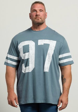 Hombre con cabello corto y barba, que lleva una camiseta azul holgada con el número blanco 97 y mangas a rayas, de pie frente a un fondo claro y sencillo.