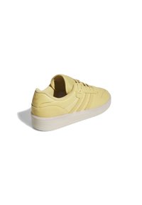 Zapatilla de cuero amarilla con detalles perforados, tres rayas laterales y una suela de goma blanca. Presenta un logotipo en relieve en el talón.