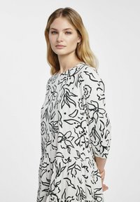 Czarna sukienka w biało-czarny wzór kwiatowy z dekoltem w łódkę, rękawami do trzech ćwiartek oraz warstwowym falbanowym wzorem. Gładka tekstura materiału.