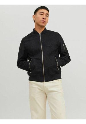 Junger Mann trägt schwarze Bomberjacke mit Reißverschluss und Taschen sowie cremefarbene Hose, steht vor schlichtem hellem Hintergrund.