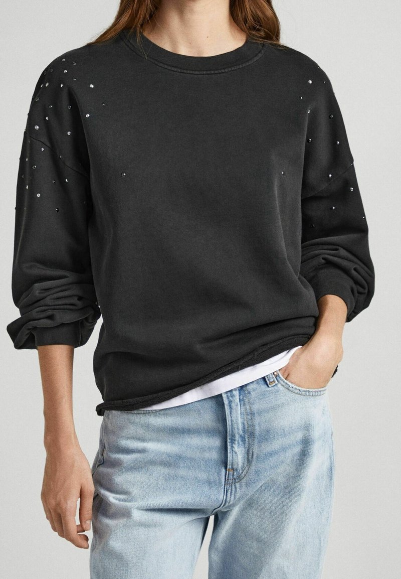 Svart sweatshirt med långa ärmar, pryd med dekorerade nitar på axlarna, avslappnad passform och synlig fåll, tillsammans med en vit t-shirt och ljusa jeans.