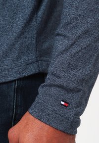 Detailní záběr na manžetu tmavě šedé košile s dlouhým rukávem, na které se u zápěstí nachází malé obdélníkové logo v červené, bílé a modré barvě, oblečené přes tmavé džíny.