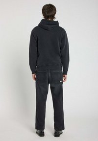 Haut à capuche noir avec capuche et poignets côtelés, associé à un jean sombre ample, comportant des poches arrière et une étiquette sur la taille.