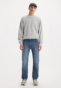 Szary sweatshirt z logo "LEVI'S", niebieskie dżinsy i szare sneakersy. Mężczyzna stoi przodem, nosząc granatową czapkę. Prosty, casualowy strój.