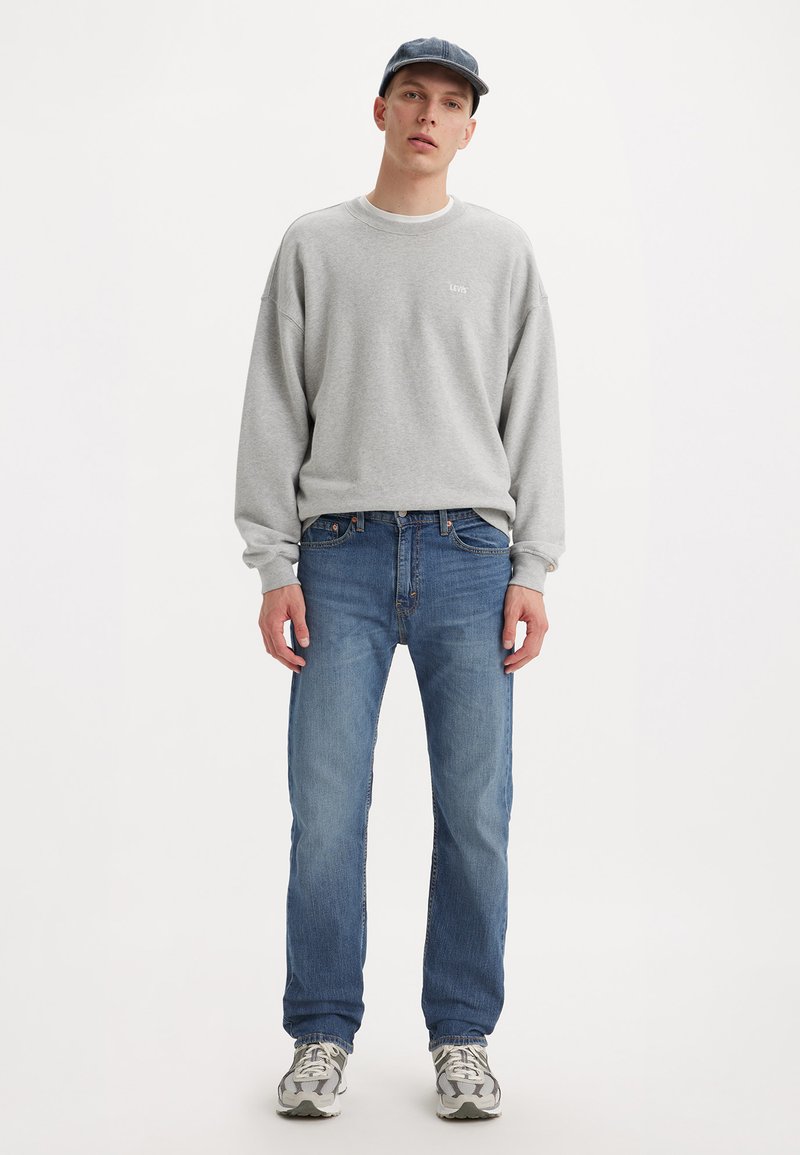 Szary sweatshirt z logo "LEVI'S", niebieskie dżinsy i szare sneakersy. Mężczyzna stoi przodem, nosząc granatową czapkę. Prosty, casualowy strój.