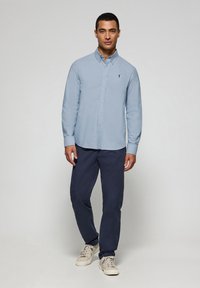 Light blue long-sleeve shirt with a button collar, featuring a small logo on the chest. Paired with navy pants and beige sneakers.