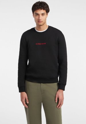 Sudadera negra de algodón con puños y dobladillo acanalados, que presenta un logo rojo "GUESS JEANS" en el pecho en una fuente estándar.
