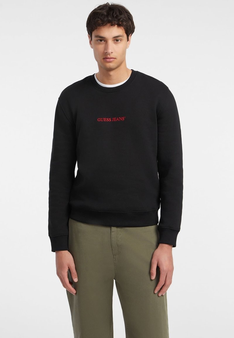 Black sweatshirt made of cotton with ribbed cuffs and hem, featuring a red "GUESS JEANS" logo across the chest in a standard font.