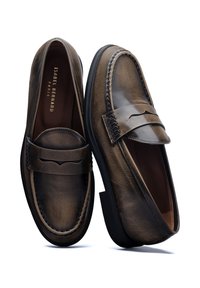 Mocassins en cuir marron avec couture visible, semelle plate et une bande décorative sur le devant. Texture lisse avec une finition polie.