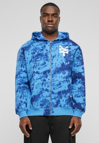 Haut à capuche zippé bleu tie-dye avec un logo blanc, poches avant et poignets côtelés. Tissu doux au toucher, présentant différentes nuances de bleu.
