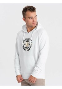 Sudadera blanca con capucha y bolsillo tipo canguro, que presenta un diseño gráfico circular de texto en negro y dorado en la parte delantera. Mangas largas.