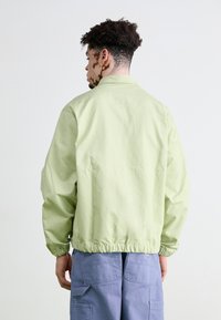 Jachetă bomber verde deschis cu manșete și tiv elastic, asortată cu pantaloni cargo bleumarin, prezentată din spate pe un fundal simplu.