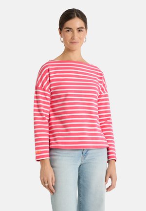 Woman with dark hair wearing pink and white striped long-sleeve shirt and light blue jeans, standing against a plain background.