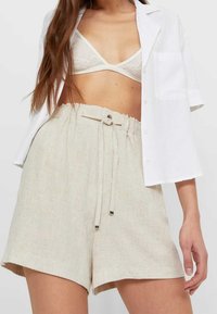 Vrouw in beige hoge taille drawstring shorts, witte kant bralette en een losse witte blouse met korte mouwen.