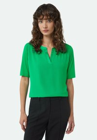 Green, short-sleeve blouse with a V-neck and smooth, lightweight fabric. Paired with black pants, showcasing a relaxed fit.