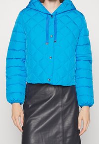 Kvinna som bär en blå vadderad kort jacka med tryckknappar och en svart läderkjol, visad från axlarna till mitten av låren.