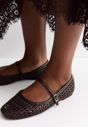 Feet wearing dark brown woven Mary Jane flats with a gold buckle strap, paired with a black lace dress hem.