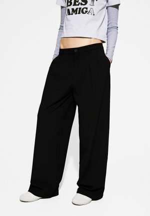 Person iført sorte bukser med brede ben, hvide sneakers og en cropped hvid top med teksten "BEST AMIGA" over en grå langærmet skjorte.