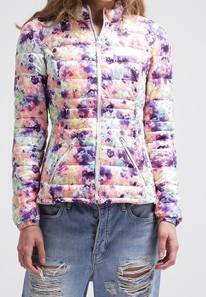 Kvinna iklädd en färgglad, blommönstrad vadderad jacka med dragkedjefickor fram och slitna blå jeans med stora hål.