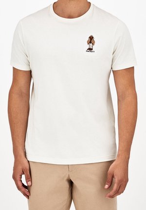 Homme portant un t-shirt blanc à manches courtes avec un petit logo d'oiseau marron et le texte "HACKETT" sur la poitrine gauche, associé à un pantalon beige.