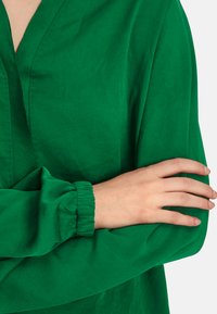 Green long-sleeve blouse with a V-neck, made of smooth fabric. Features gathered sleeves and a relaxed fit.