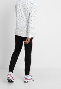 Persoon die een wit longsleeve shirt met zwarte stippen, zwarte broek met witte en rode zijstrepen, en witte sportschoenen draagt.
