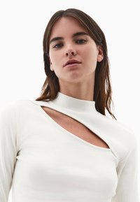 Witte geribbelde langearmige top met een hoge hals en een opvallende uitsparing aan de linkerkant, die de gladde stoftextuur benadrukt.