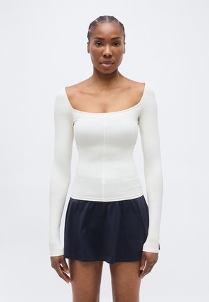 Žena s pletenými vlasmi, oblečená v priliehavom bielom topu s odhalenými ramenami a dlhými rukávmi, a tmavomodrej sukni, stojaca pred jednoduchým pozadím.