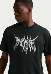 Άνδρας που φοράει μαύρο T-shirt με αφηρημένο μεταλλικό ασημένιο σχέδιο γραφικών στο στήθος, κοιτάζοντας προς τα αριστερά του, μπροστά σε απλό φόντο.