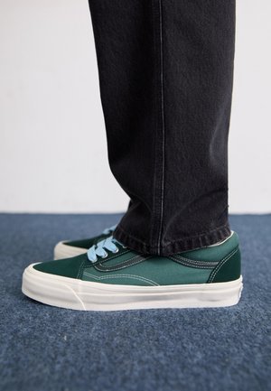 Zöld vászon sportcipő kék fűzőkkel, fehér gumitalpakkal és kontrasztos varrással. Fekete farmernadrággal viselve, oldalnézetből.