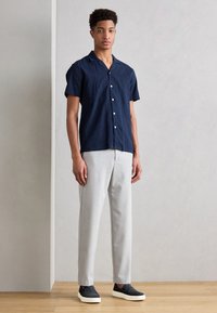 Camicia navy a maniche corte con bottoni bianchi, abbinata a pantaloni grigi chiari e scarpe scure slip-on. Texture lisce e vestibilità casual.