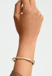 Bracciale in metallo color oro con un design liscio e curvo e accenti testurizzati, poggiato su una mano. Lucentezza sottile e stile minimalista.