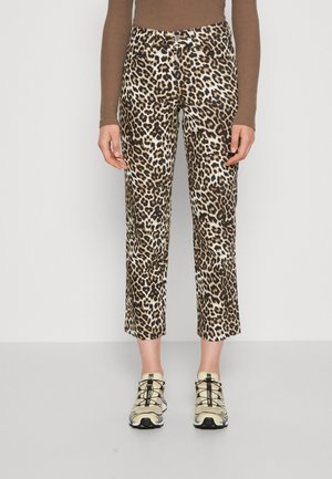 Osoba nosząca skrócone spodnie w panterkę, brązową bluzkę z długim rękawem oraz beżowe trampki z czarnymi sznurowadłami, stojąca na tle jednolitego tła.