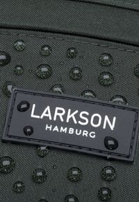 Zöld textúrájú szövet vízcseppekkel és egy fekete téglalap alakú címkével, amelyen vastag fehér betűkkel olvasható a "LARKSON HAMBURG" felirat.