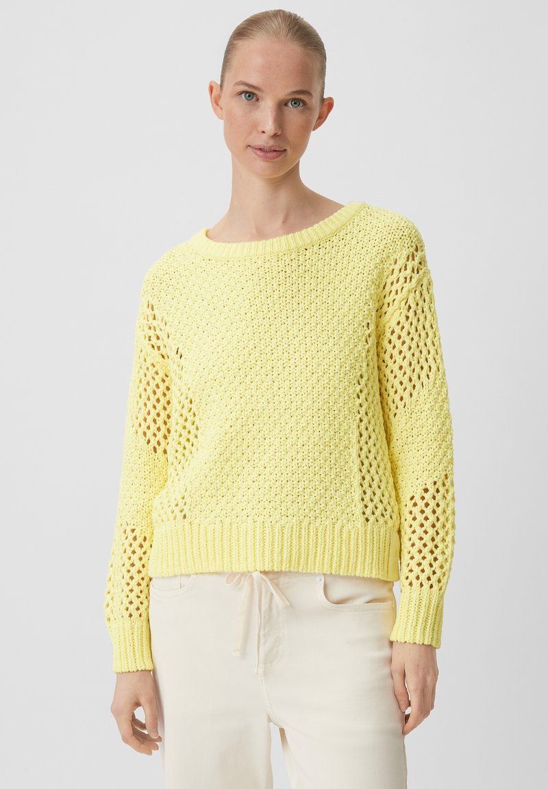 Gelb gestrickter Pullover mit Lochmuster, weitem Ausschnitt und gerippten Bündchen. Leichte Textur, cropped Design, kombiniert mit hellen Hosen.